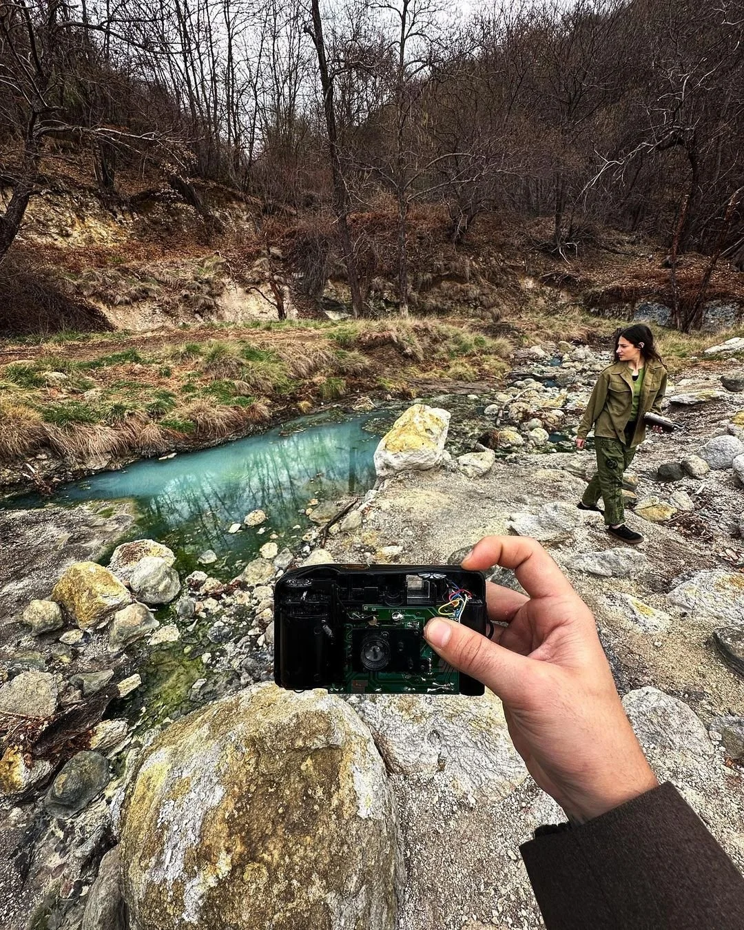 A person holding a camera in a hand taking a picture of a woman in a green jacket walking near a small river or creek in a wooded area.