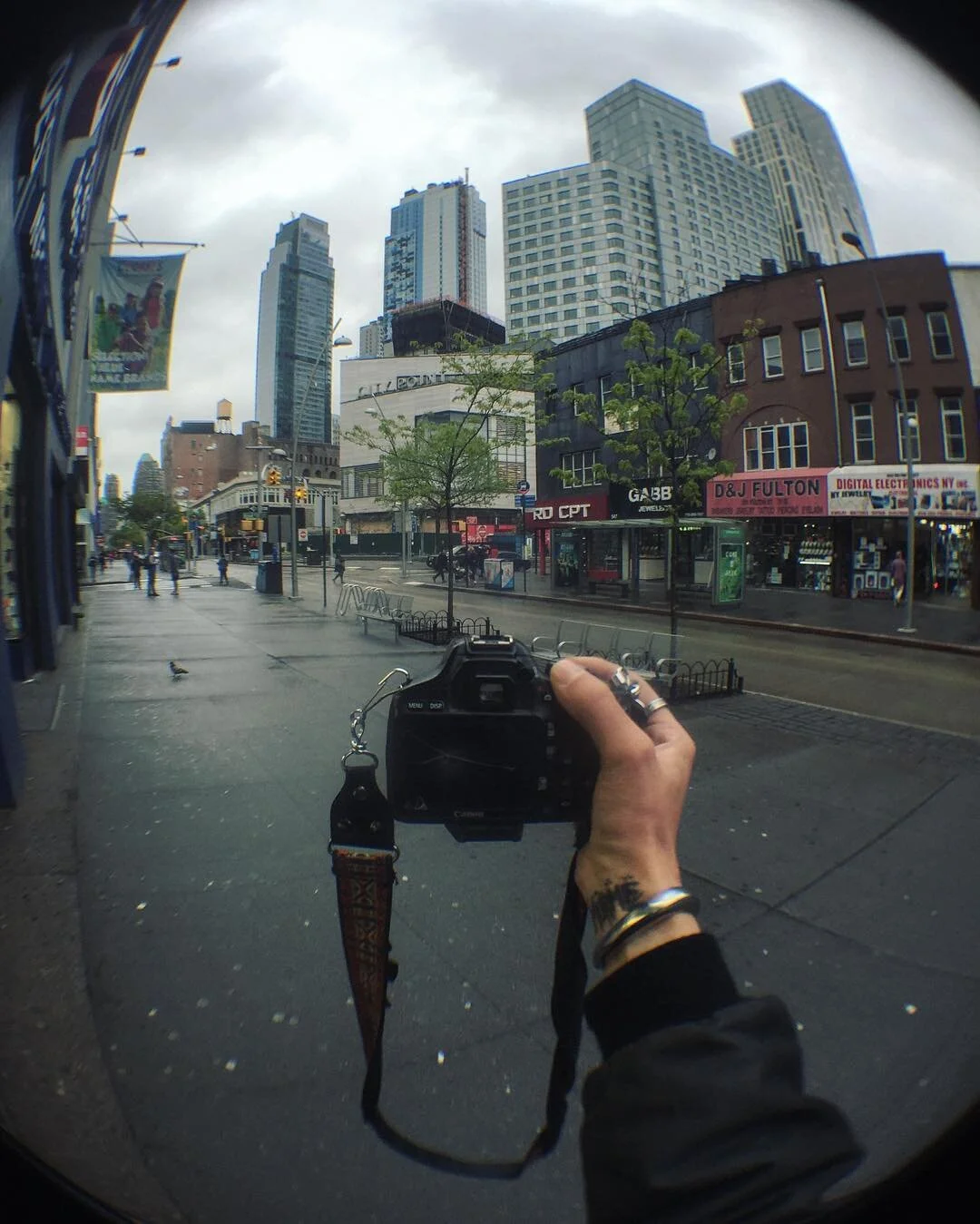 City street scene with tall buildings, storefronts, and a person holding a camera in front of a mirror, capturing the surroundings. Overcast sky with some pedestrians walking.