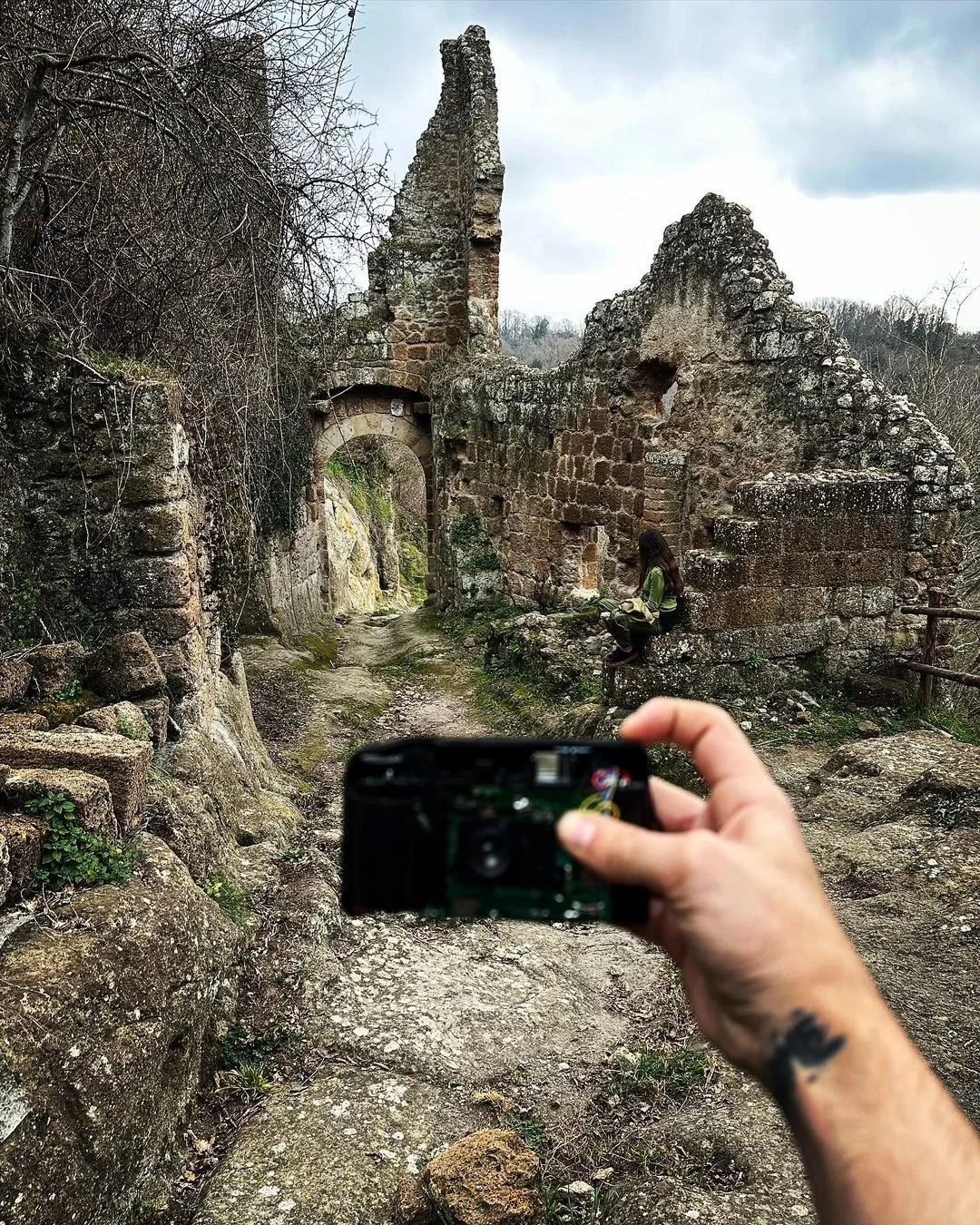 A person is taking a photo of a young woman sitting on a stone ledge among the ruins of an old stone structure in a wooded area, with cloudy sky overhead.