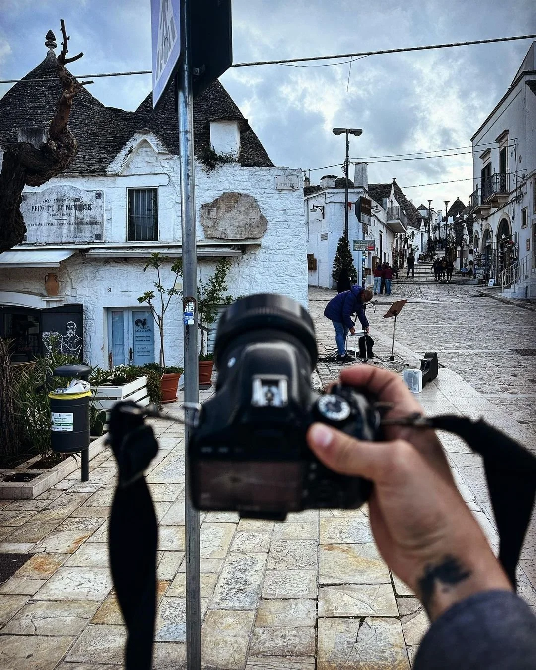 Street scene in a European town with white buildings, cobblestone street, and people walking, while a person takes a photo of street performers in the foreground.