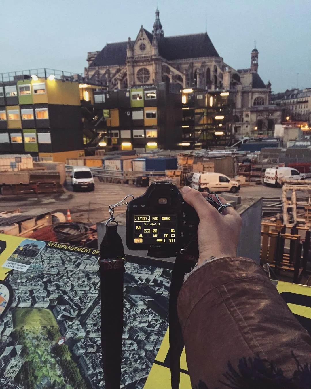 Person taking a photo of a historic church building with a camera. The scene appears to be a construction site with forklifts, trucks, and building materials, in front of colorful modern apartments.