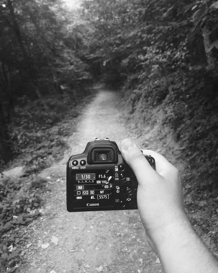 A person holding a Canon camera, taking a photo down a forest trail with trees on both sides.