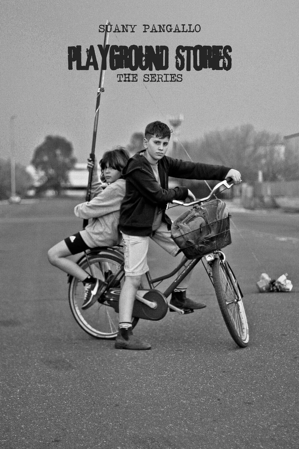 Two children, a boy and a girl, are riding a bicycle together in a street, with the girl sitting on the back and holding a pole, and the boy riding and steering in the front. The image is in black and white.