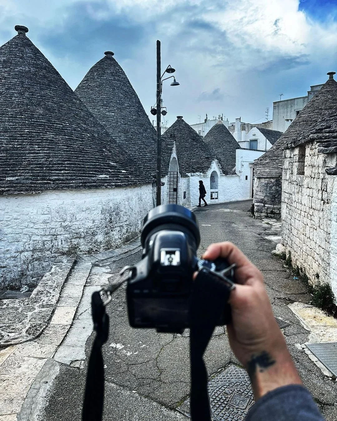 Person holding a DSLR camera taking a picture of a narrow street with traditional white stone houses and conical roofs in a European town, with a person walking in the background.