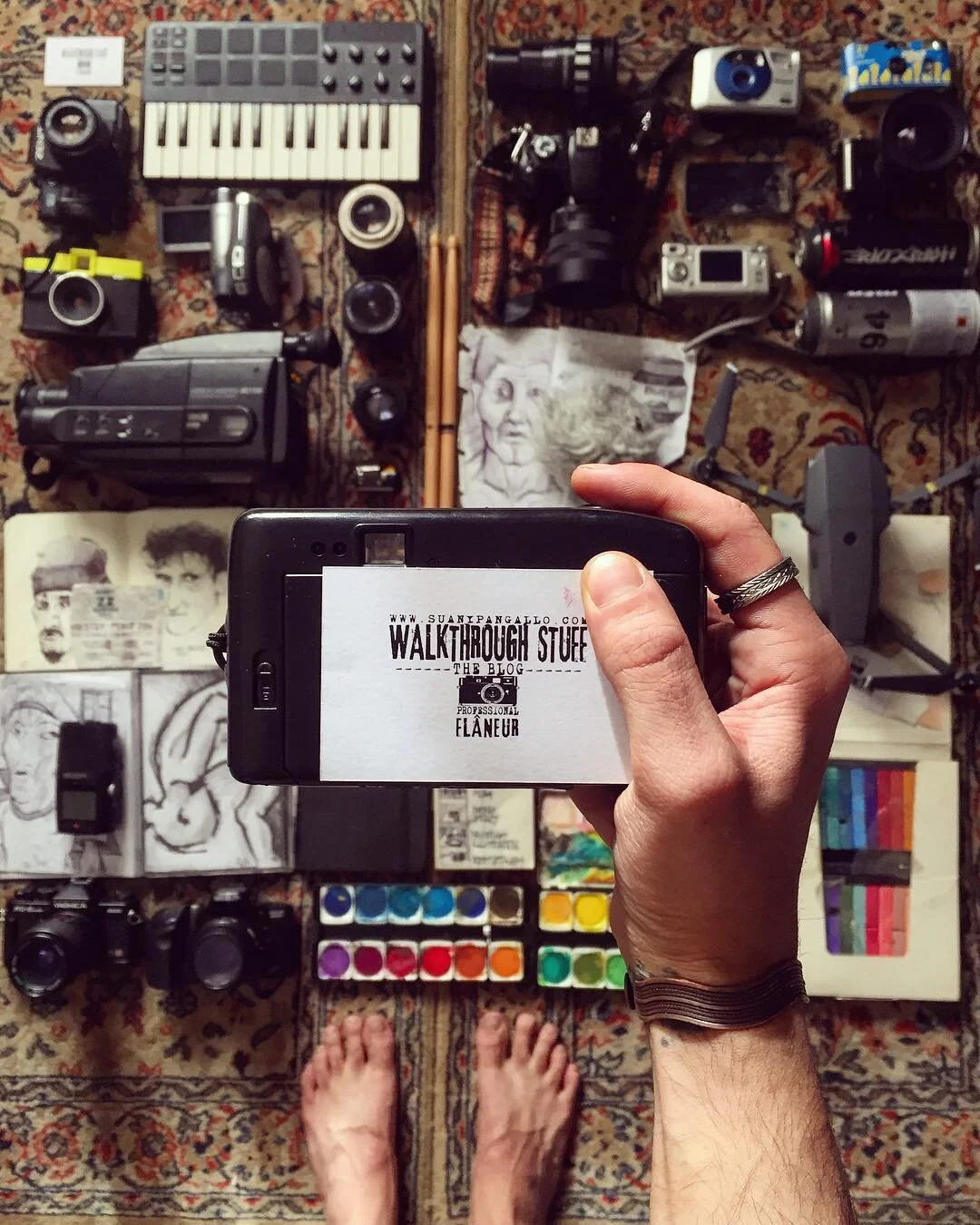 A person taking a photo of a collection of art supplies, cameras, audio equipment, and sketches arranged on a patterned rug. The person's feet are visible at the bottom of the image.