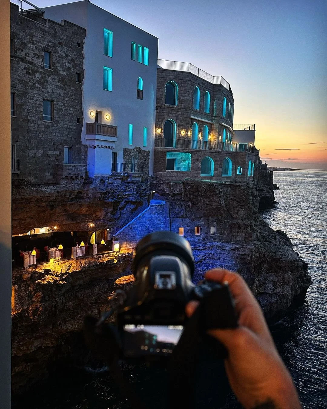 A person takes a photo of a building with glowing blue windows on a rocky cliff at sunset, overlooking the ocean.