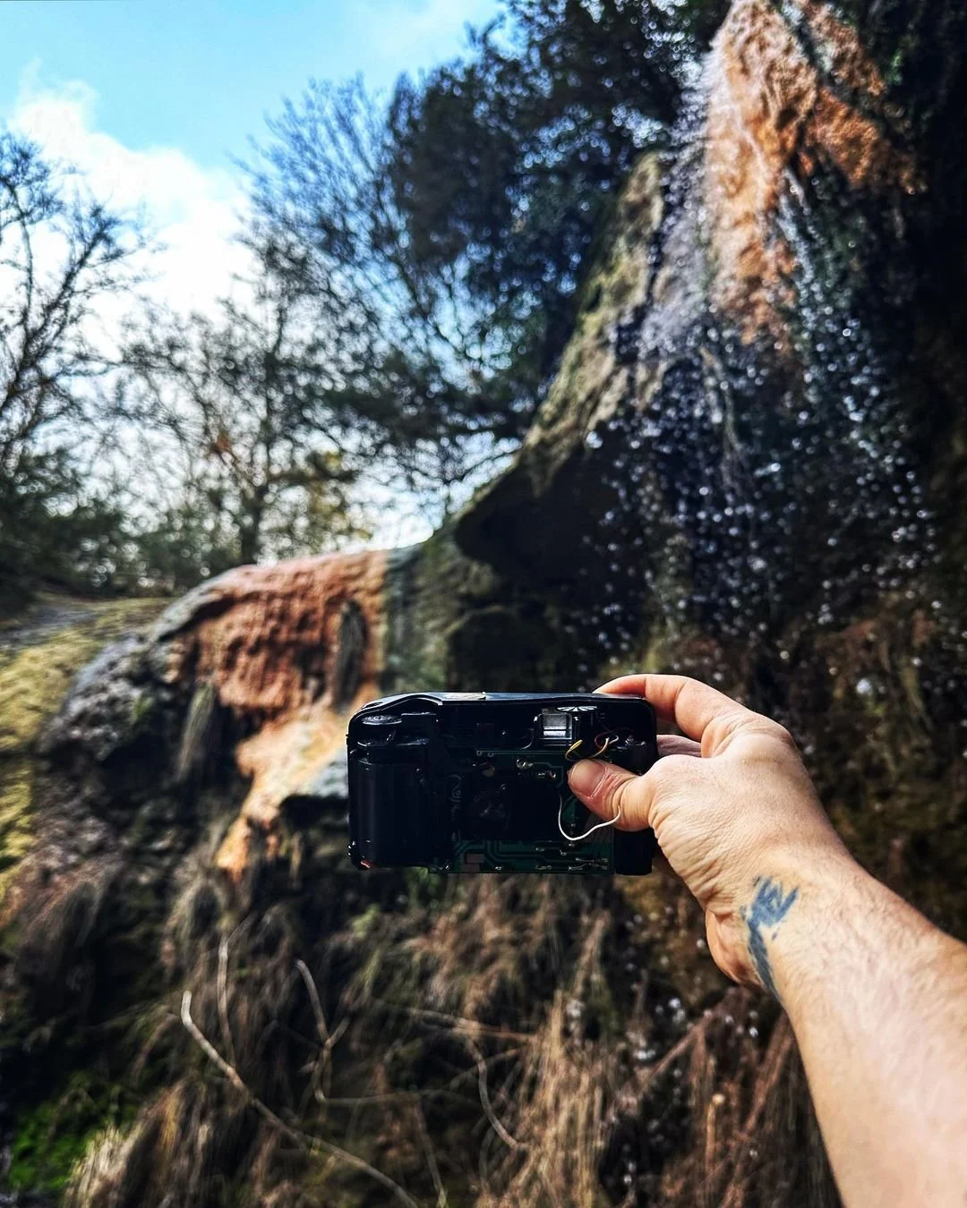 Person holding a camera taking a photo of a waterfall in a forested area with trees and blue sky in the background.