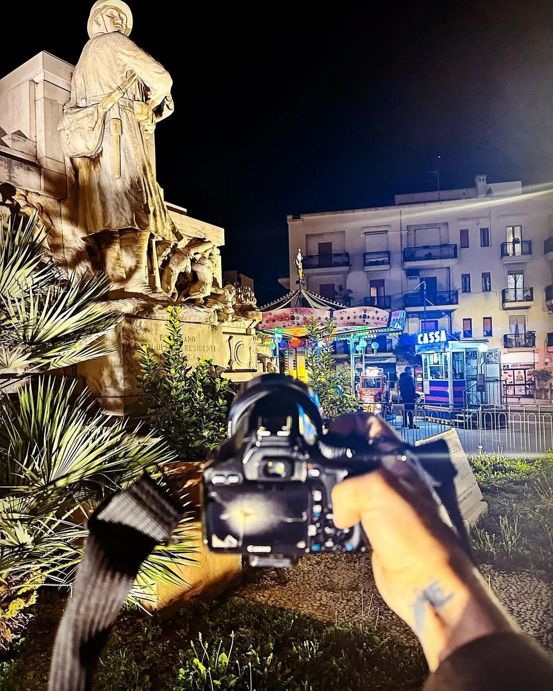 A person taking a photo of a monument at night with a camera, with a colorful amusement park in the background.