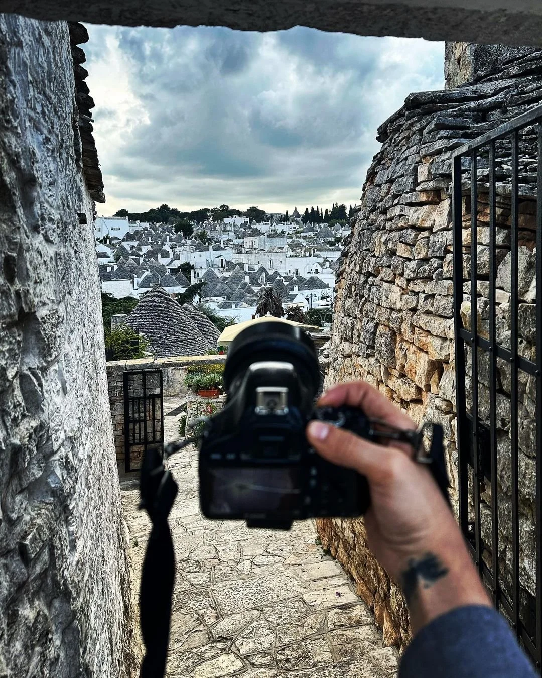 Photo of a person taking a picture of a townscape with a DSLR camera, framed by stone walls and an iron gate, showing white buildings and rooftops under cloudy sky.