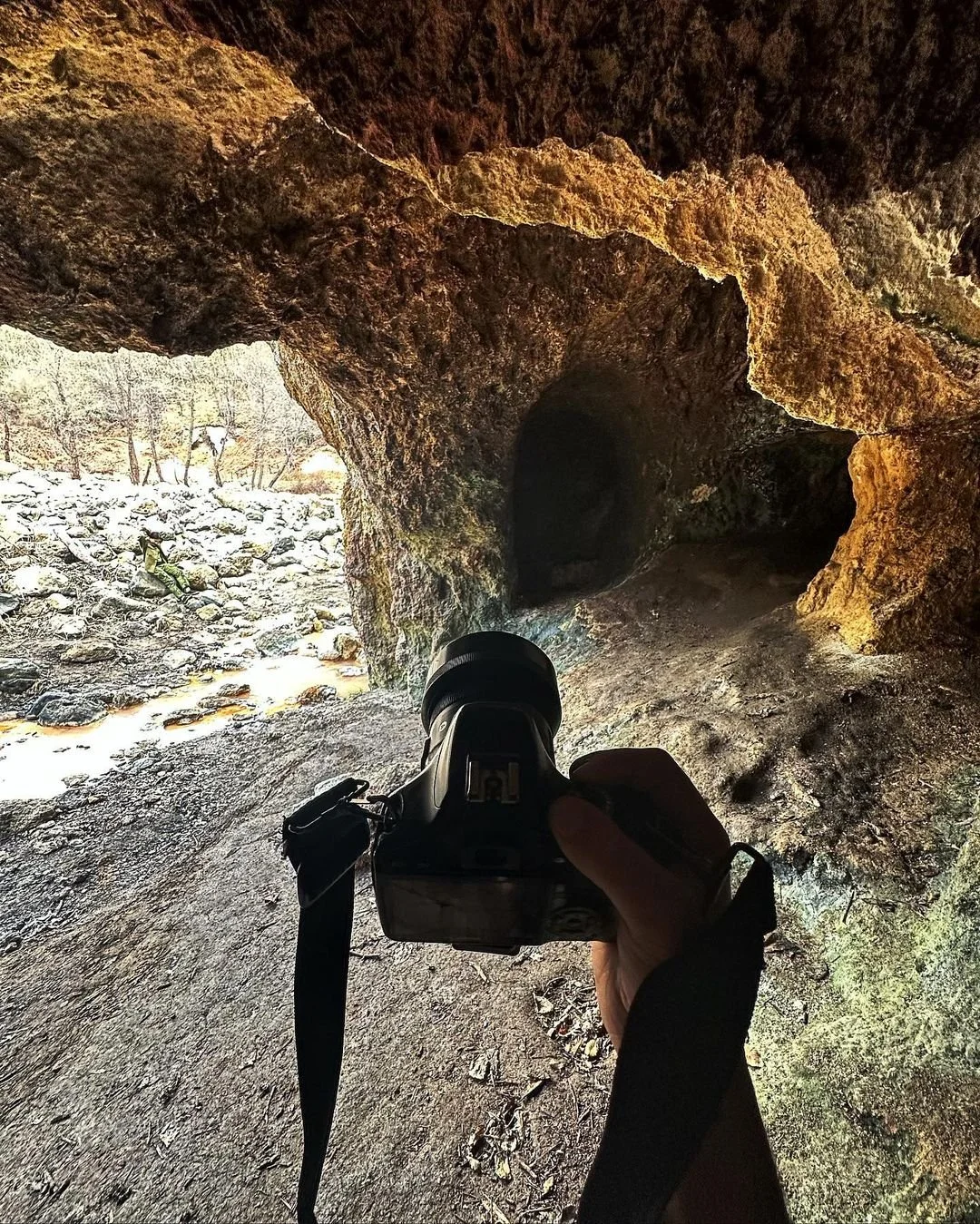 Person holding a camera inside a cave, with another person walking on rocky ground outside in the background.
