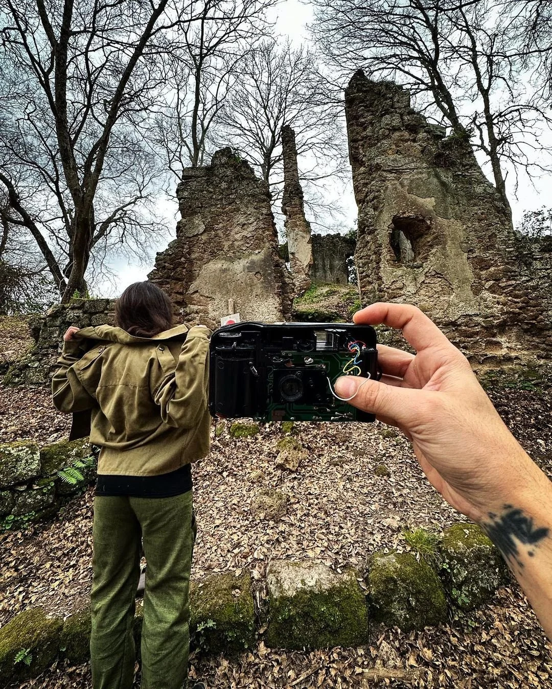 A person with a tattoo on their wrist holding a camera captures a woman in a green jacket looking at the ruins of an old stone structure in a wooded area. The ruins are surrounded by trees with bare branches and fallen leaves on the ground.