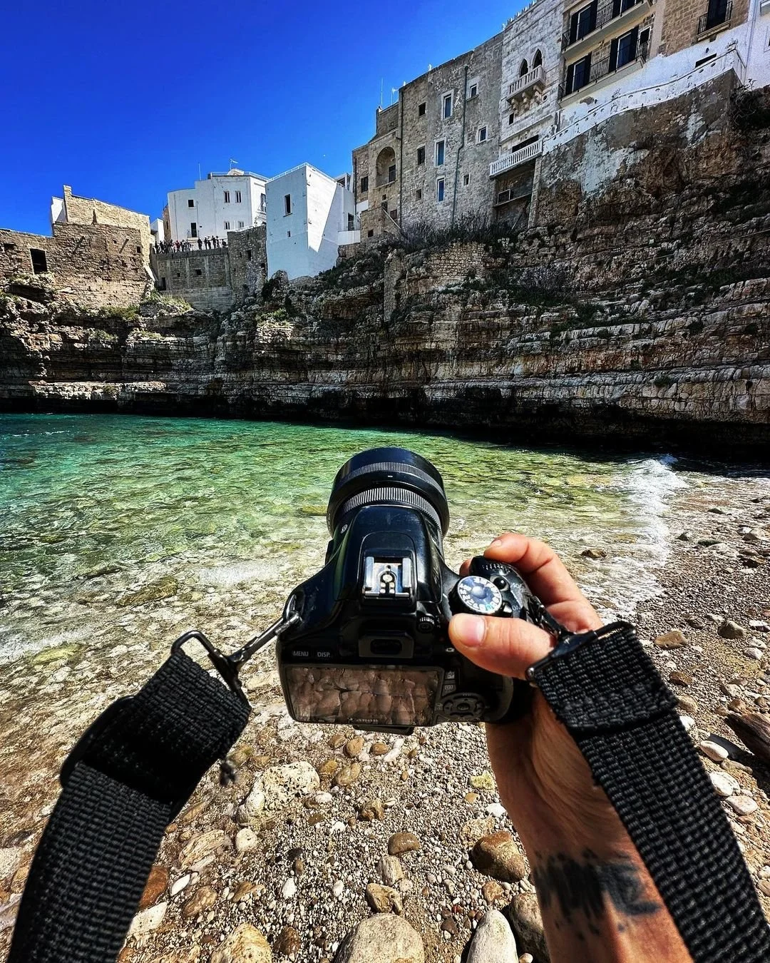 A person holding a camera taking a photo of colorful buildings along a rocky shoreline with green water and a clear blue sky.