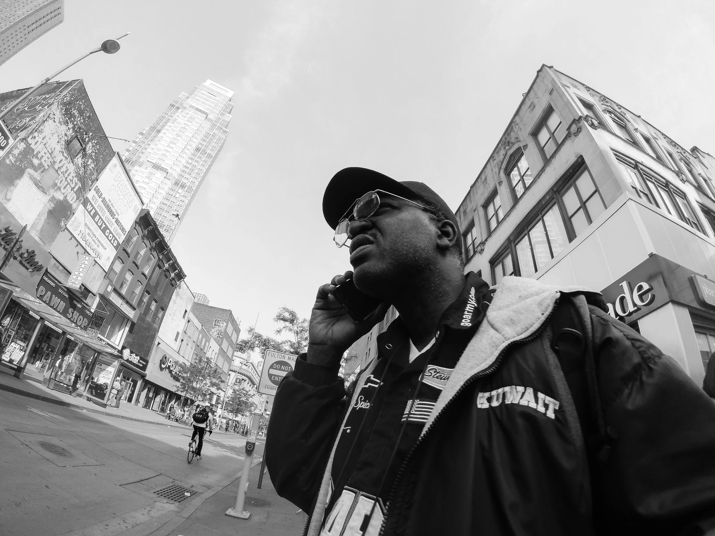 A black and white photo captures a man wearing a cap and glasses, talking on a cell phone while walking in an urban area. The background features tall buildings, billboards, a cyclist, and other pedestrians.