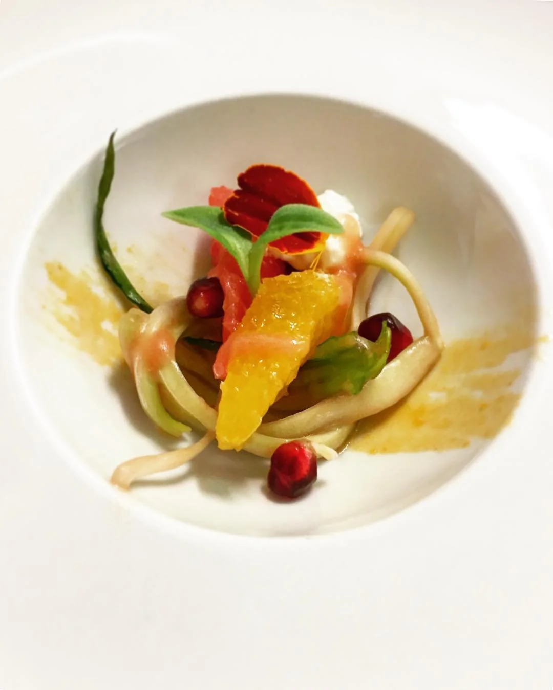 A plated gourmet dish with spaghetti, garnished with citrus segment, green leaves, red edible flowers, and pomegranate seeds.
