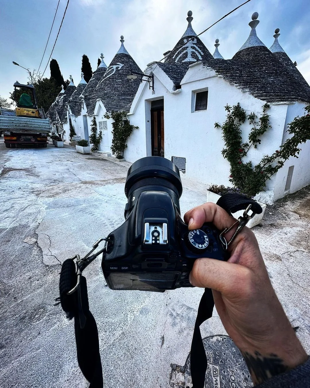 Photograph of a person holding a camera, aiming at white houses with conical roofs and climbing plants on a narrow street.