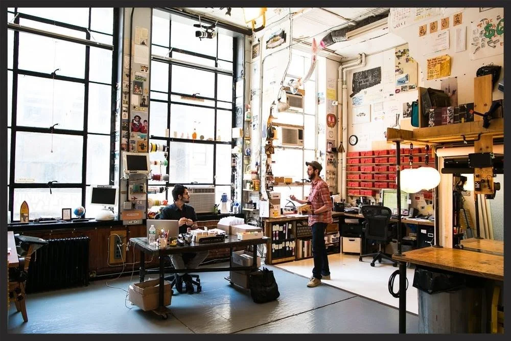 A spacious, well-lit urban workshop or studio with large windows, featuring two men engaged in conversation. One man is seated at a cluttered desk using a laptop, while the other stands holding a notebook or tablet. The room is filled with various to