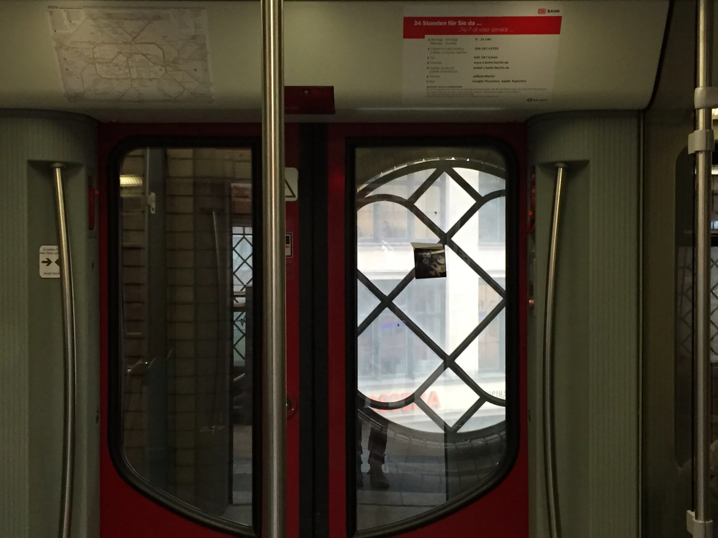Interior view of a train door with a glass window, metal poles, and a red border. Outside the window, a person is visible walking on the street.