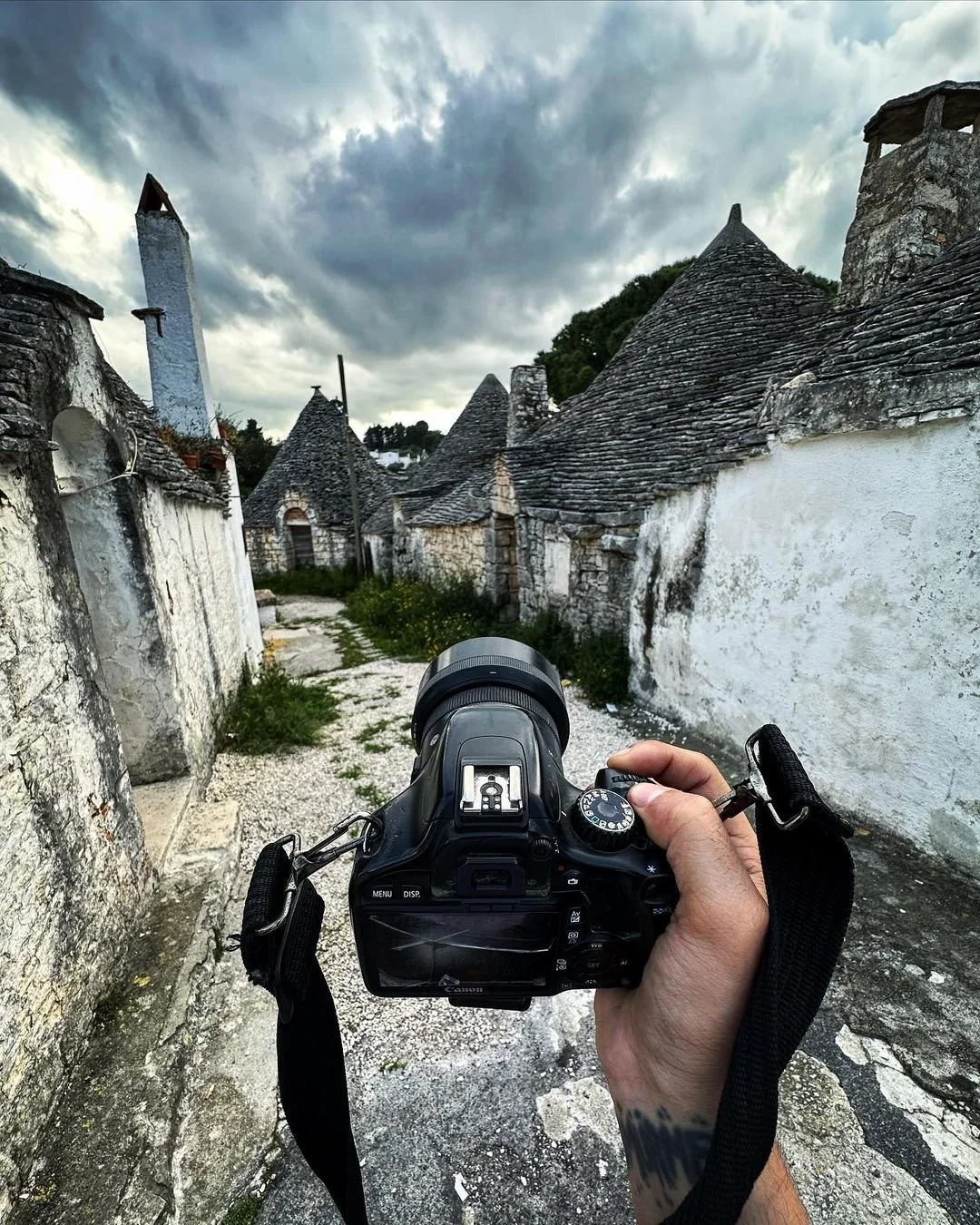 A person holding a camera takes a photo of a historic village with white stone buildings and conical roofs under a cloudy sky.