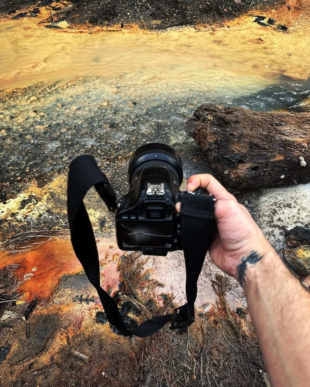 A hand holding a camera aimed at a natural hot spring or geothermal pool with colorful mineral deposits and a large piece of dark, textured wood nearby.