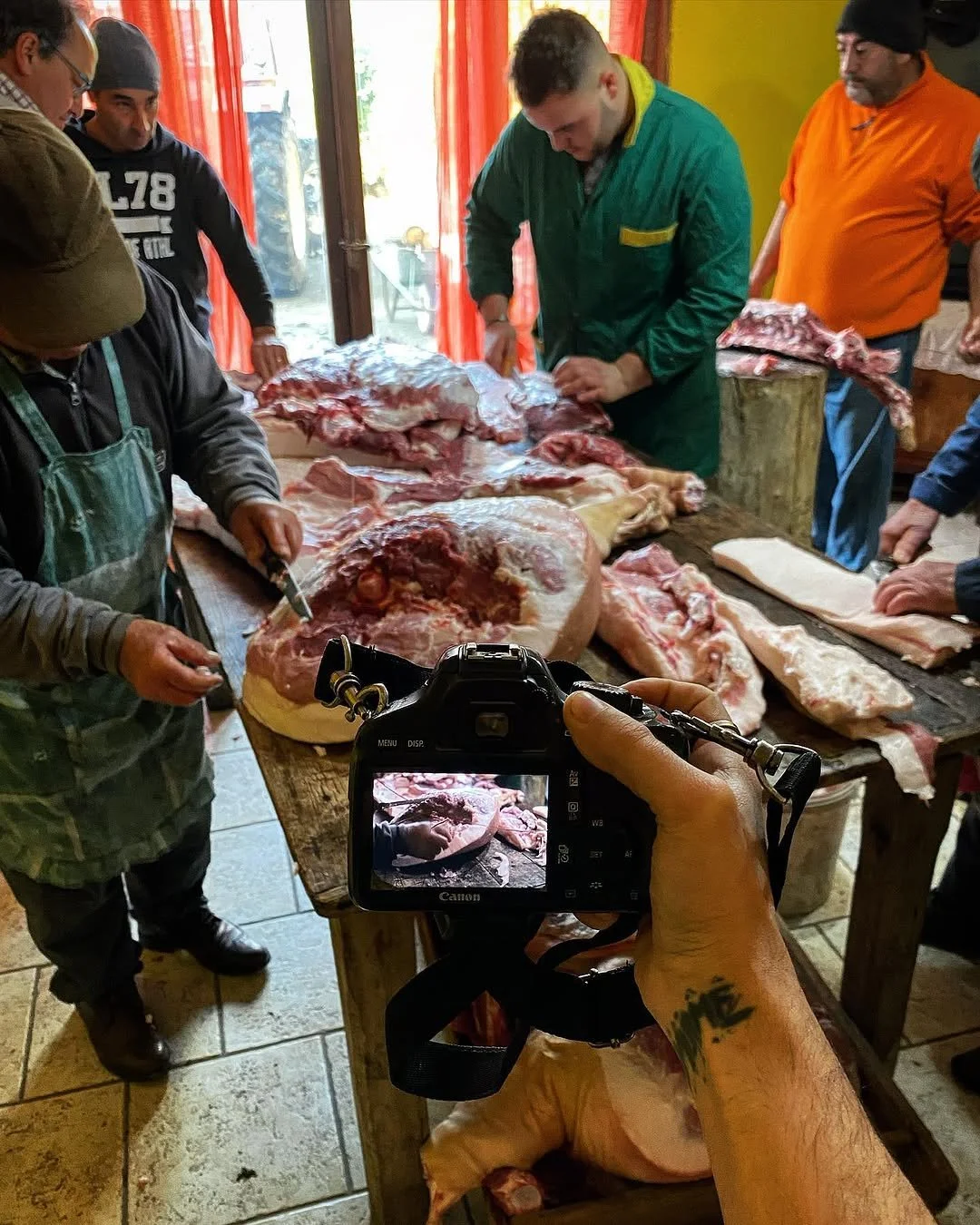 People butchering and preparing a large piece of raw meat in a room with red curtains. A person in the foreground is taking a photo of the scene with a camera.