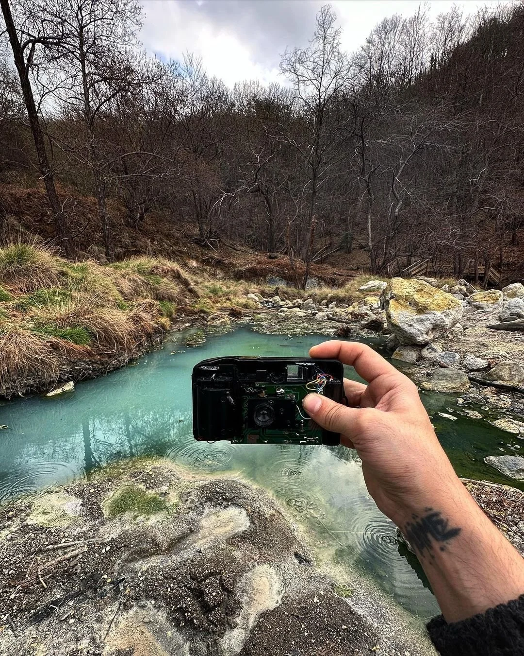 A person holding a camera over a small creek or river in a natural landscape with leafless trees and rocks.