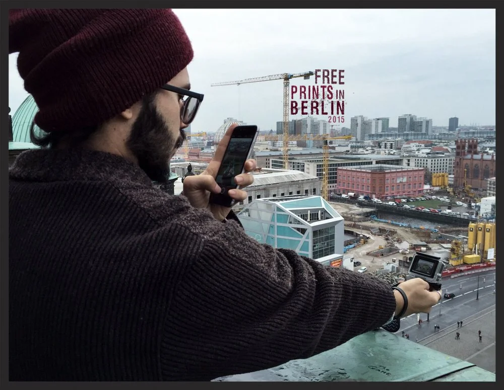 A man with glasses and a beard, wearing a maroon beanie and dark sweater, taking a photo with a camera and phone on a rooftop overlooking a cityscape with construction cranes, buildings, and a sign that reads 'FREE PRINTS IN BERLIN 2015'.