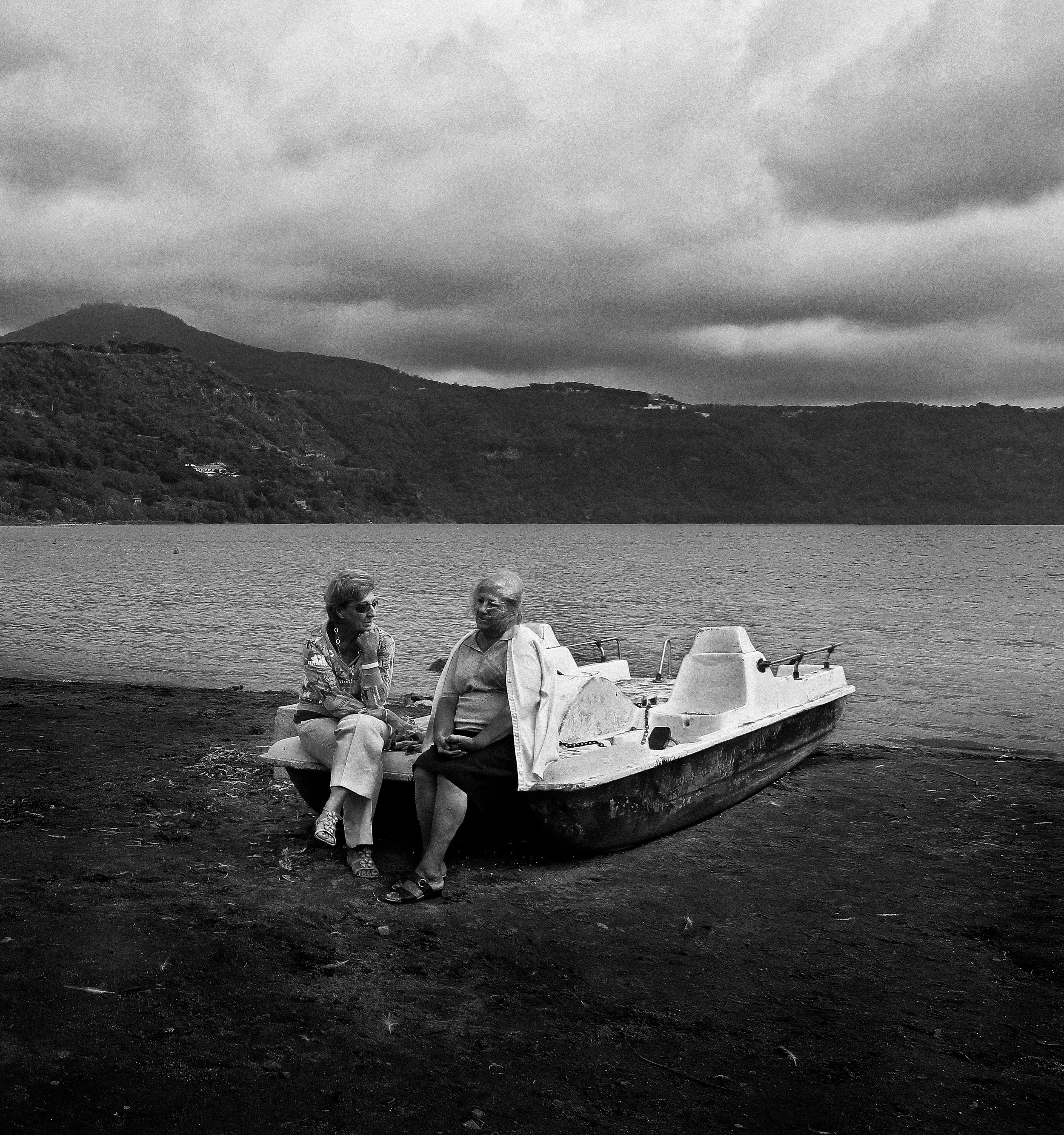Two women sit on a dock near a body of water with a mountain and cloudy sky in the background.
