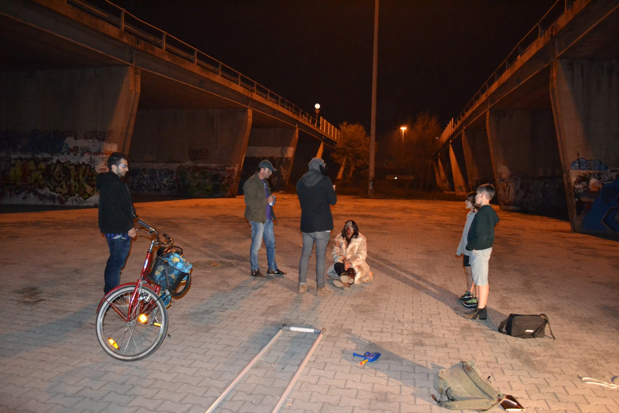 A group of five people, including children, are gathered on a paved area underneath a bridge at night. One person is sitting on the ground while others stand around them, some holding drinks. A bicycle is in the foreground, and there are tools and eq