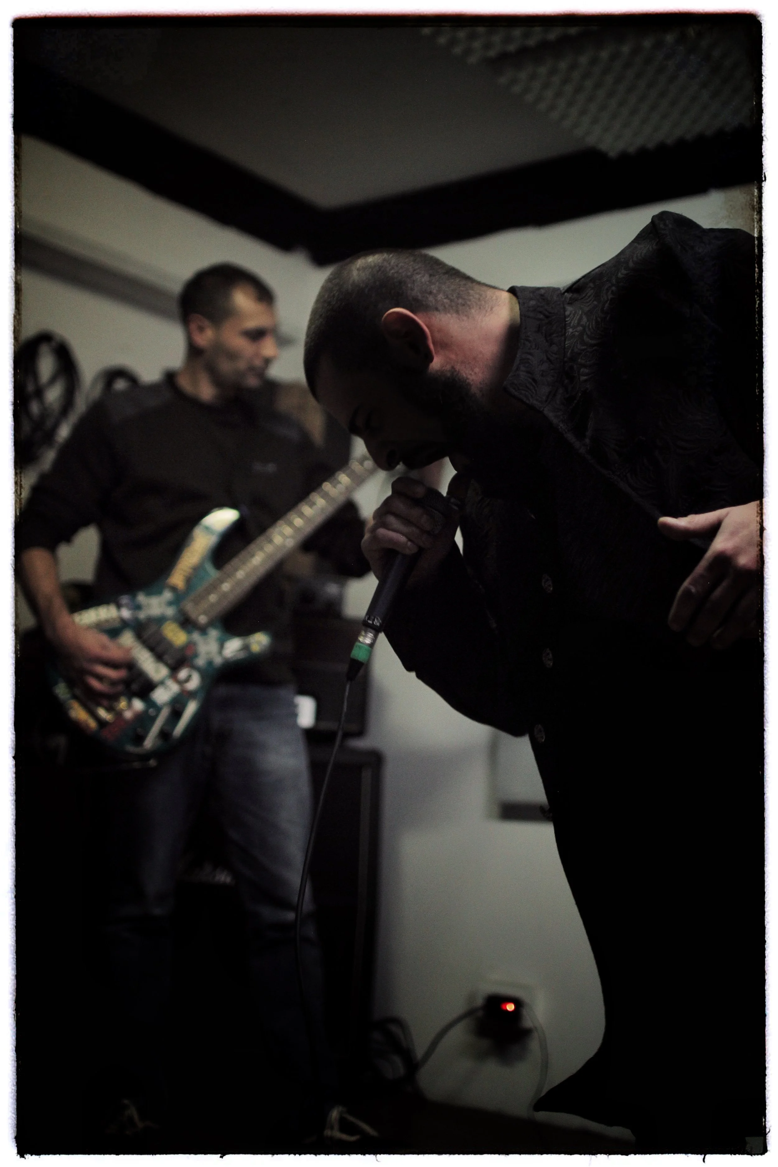 Two men are in a music studio; one is playing an electric guitar and the other is singing into a microphone with a cord, with a dark background and audio equipment.