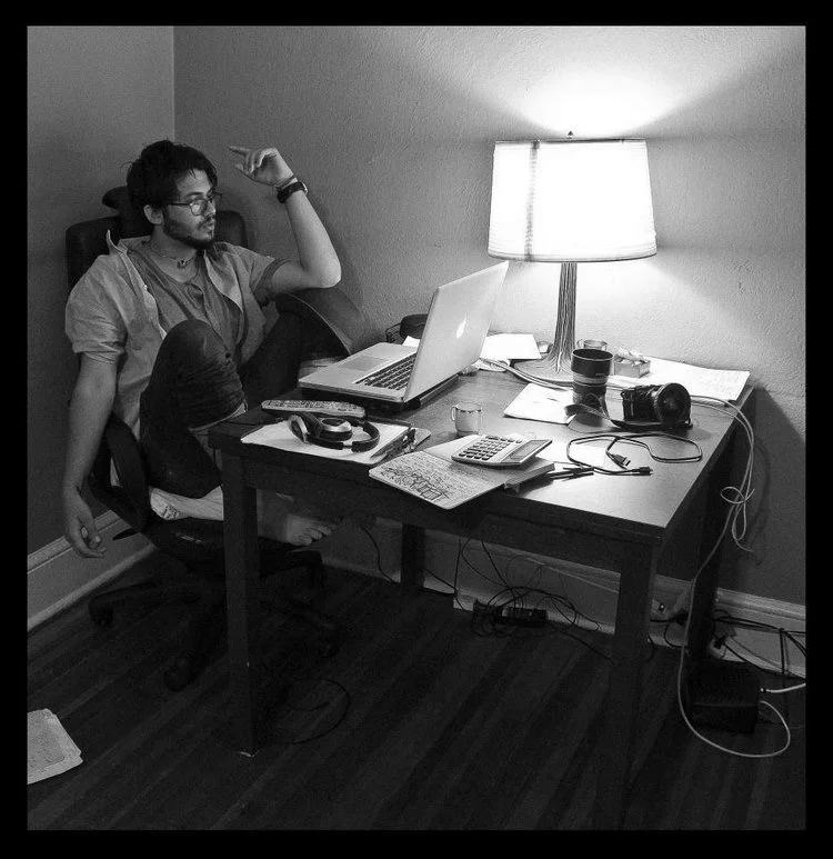 A young man with glasses and a beard sitting at a cluttered desk with a laptop, headphones, camera, calculator, and papers, in a dimly lit room with a lamp.