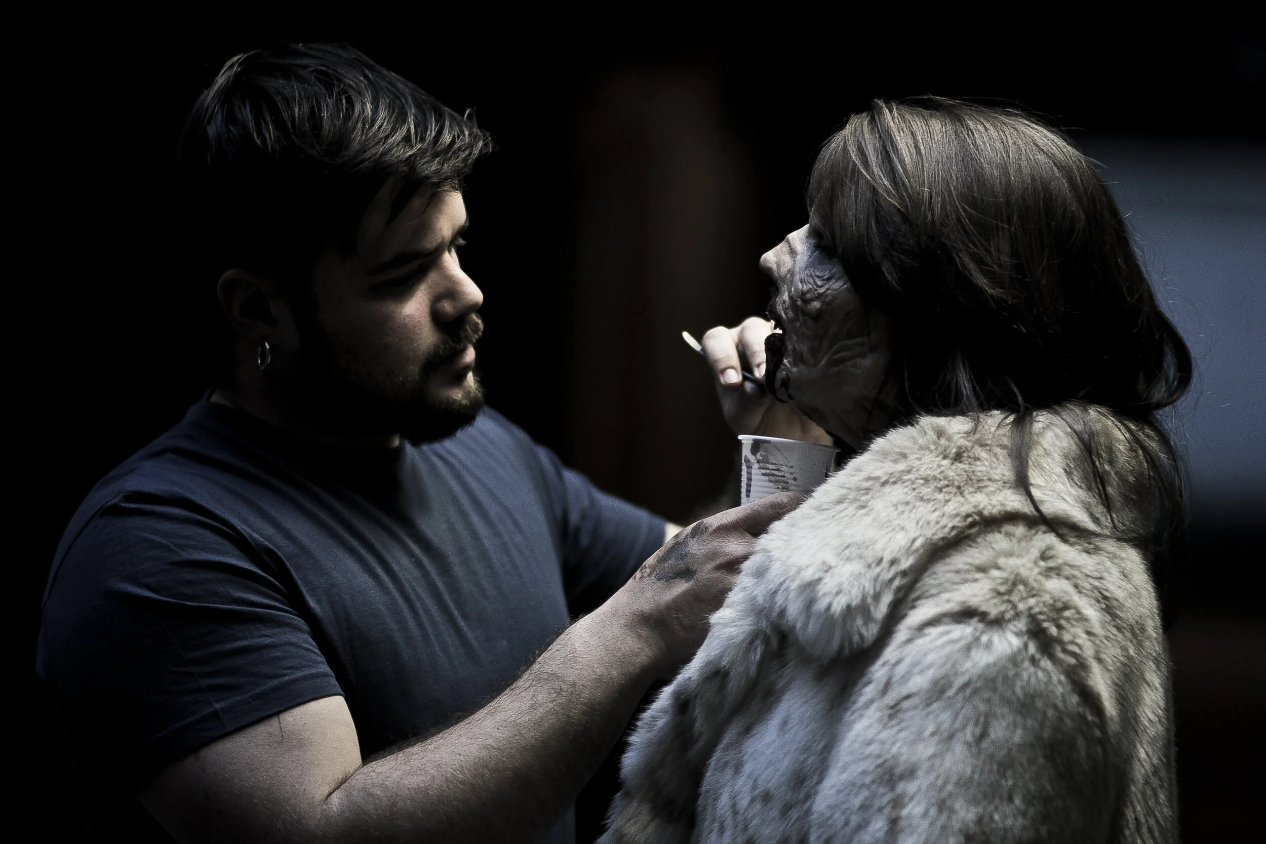 A man with dark hair and a beard feeds a large wolf with long gray hair and a white coat, holding a cup in his hand.