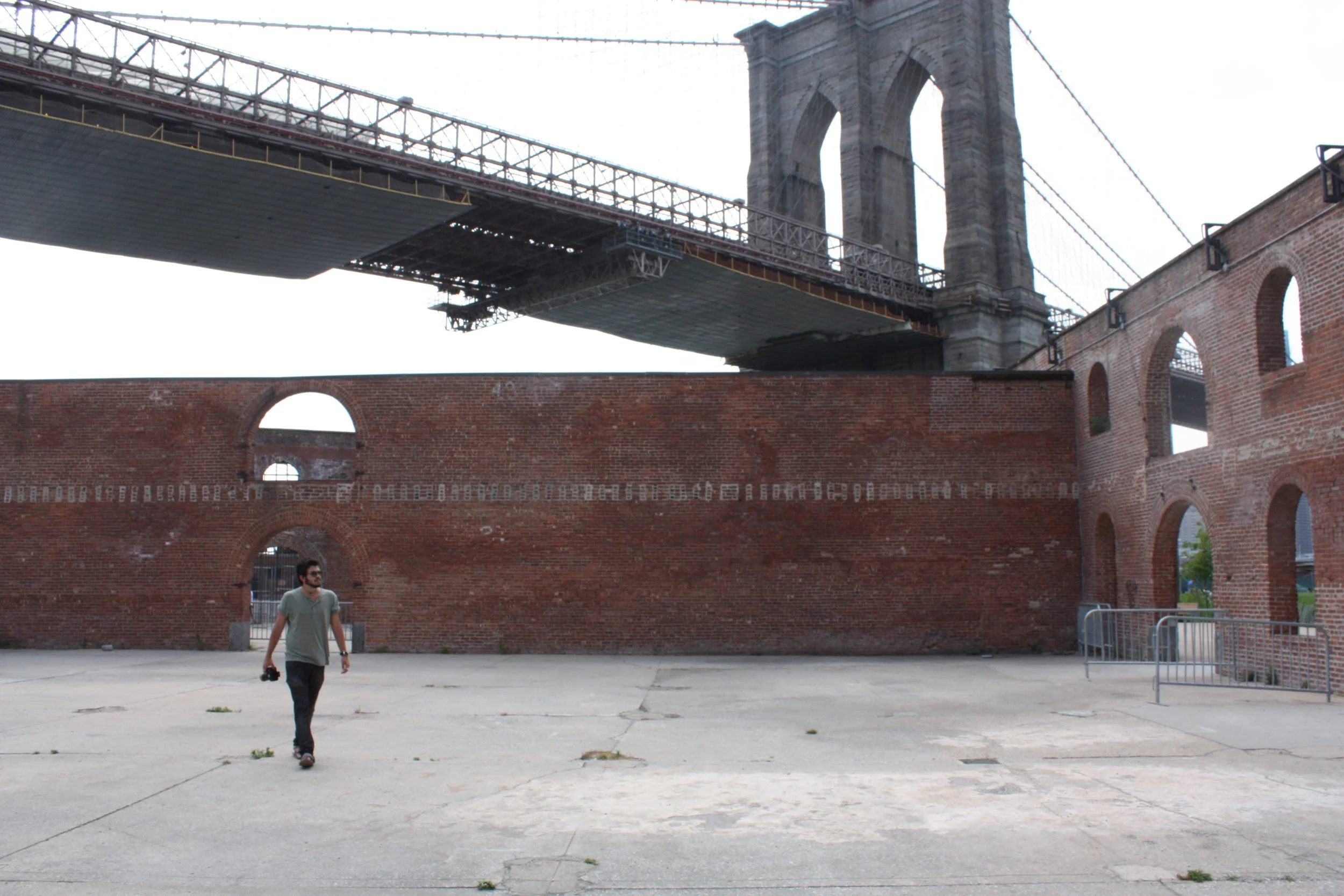 View of the Brooklyn Bridge's towers and suspension cables, with a man walking in front on an open paved area beneath a brick wall and ruins.