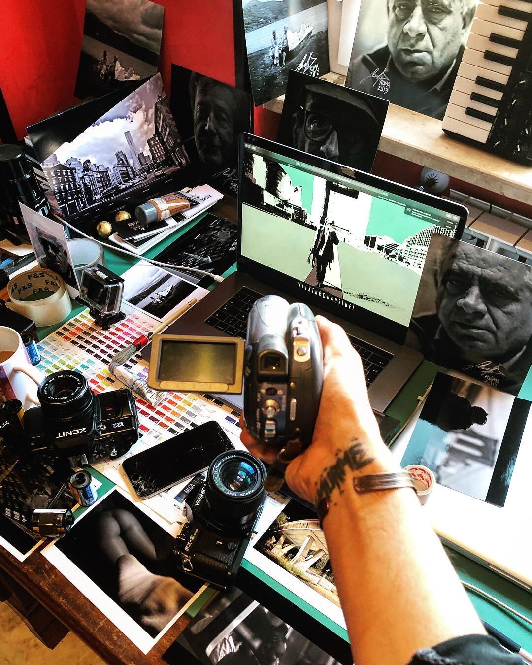 A cluttered workspace with photography equipment, printed photos, a laptop displaying a photo editing program, and portrait posters of a man on the desk and around the room. A hand is holding a camera above the workspace.