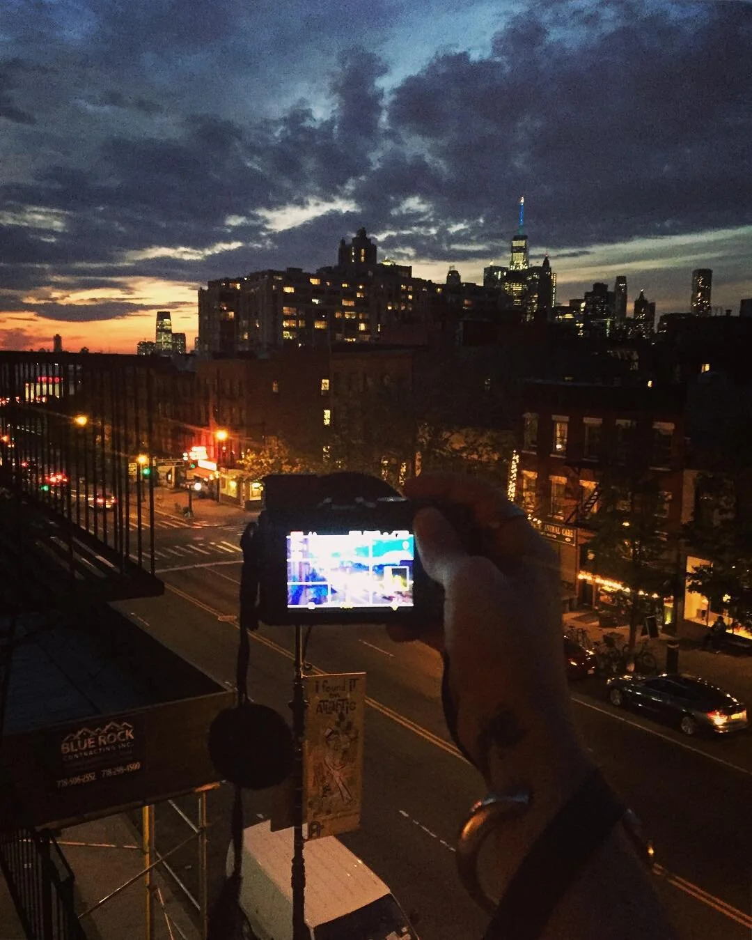 A person holding a camera, capturing a city skyline at dusk with a partly cloudy sky, tall buildings, and city lights illuminating the streets below.