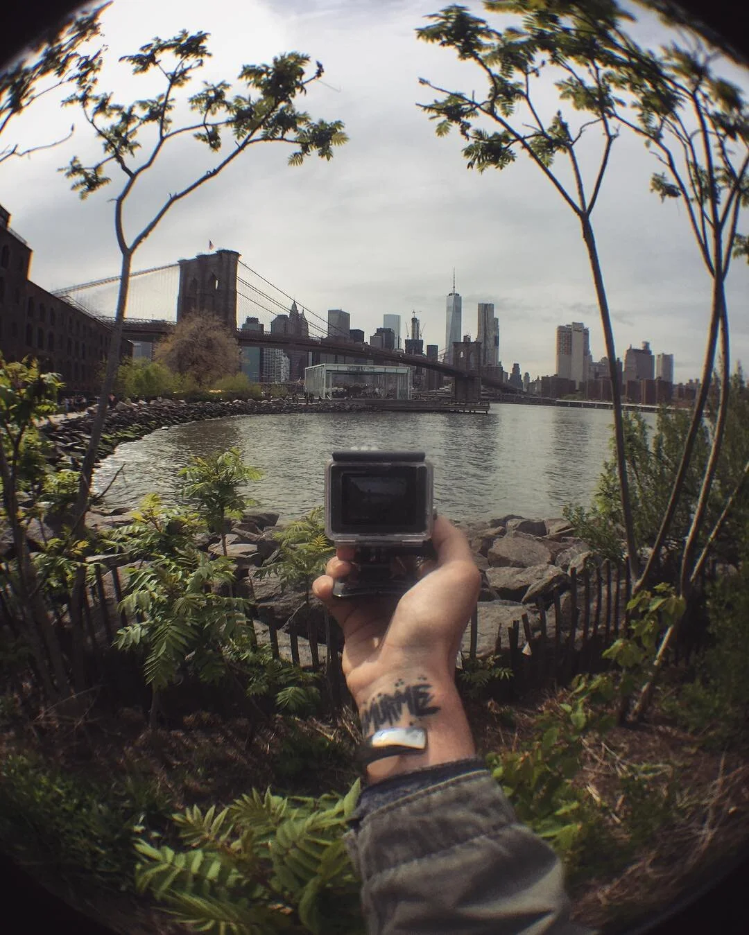 A person holding a camera taking a photo of New York City skyline across the river, with Brooklyn Bridge visible, framed by branches and greenery.