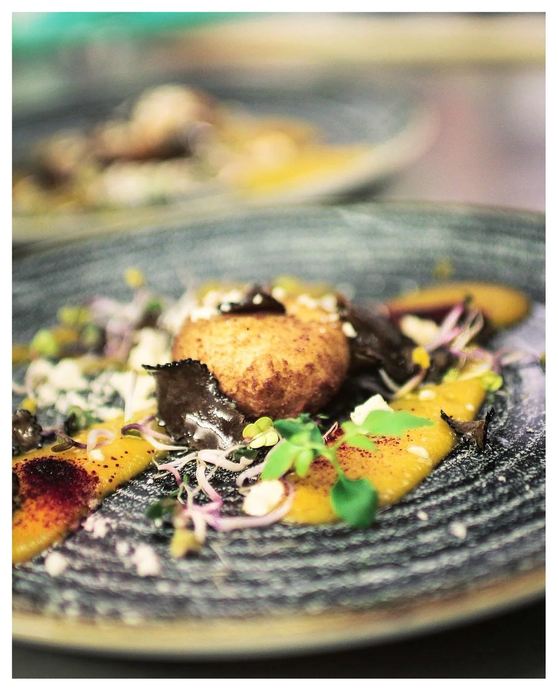 Close-up of a gourmet dish featuring a seared scallop, microgreens, colorful sauces, and edible flowers on a textured black plate.