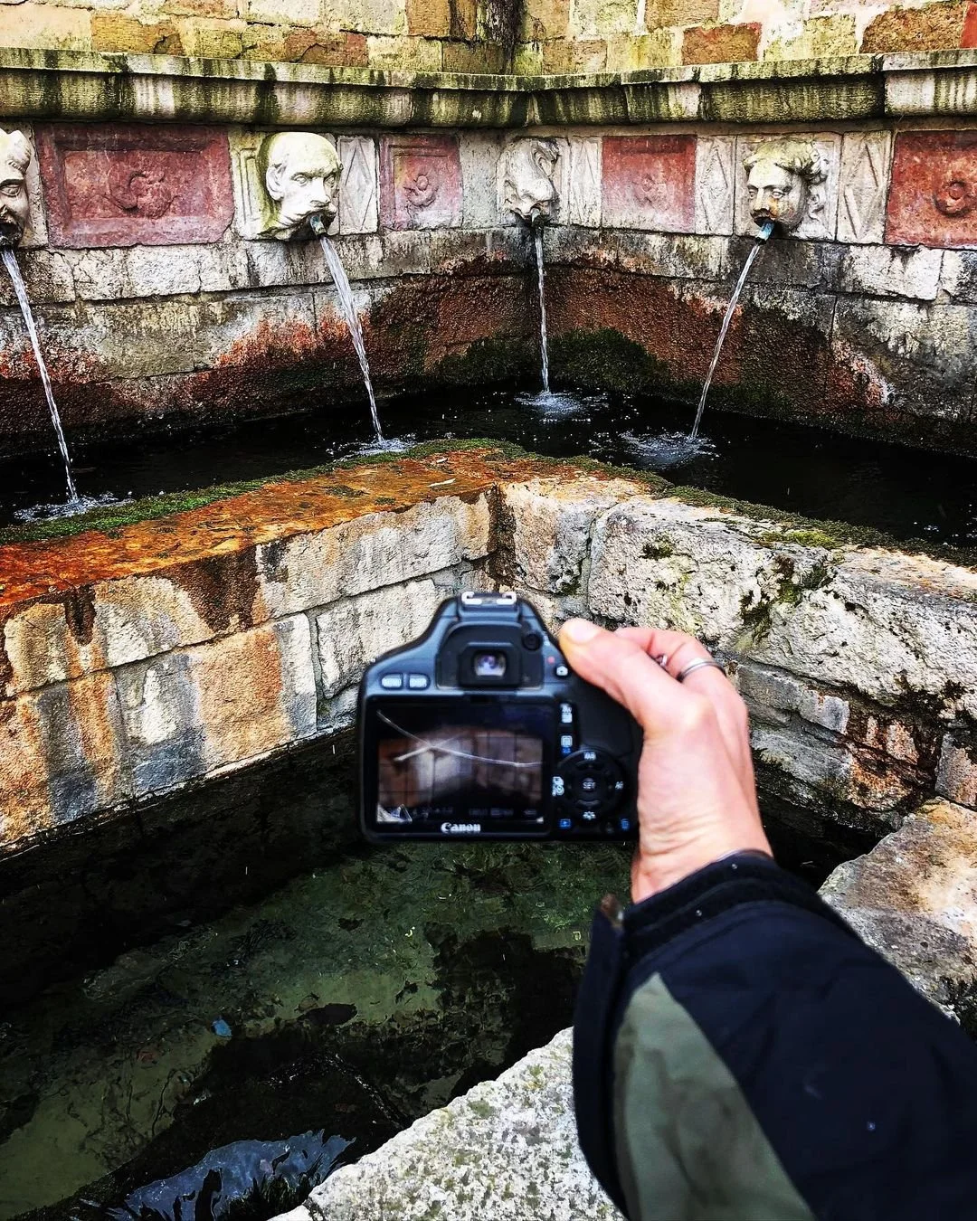 A person taking a photograph of a stone fountain with three face sculptures, each pouring water from their mouths, in an outdoor setting.