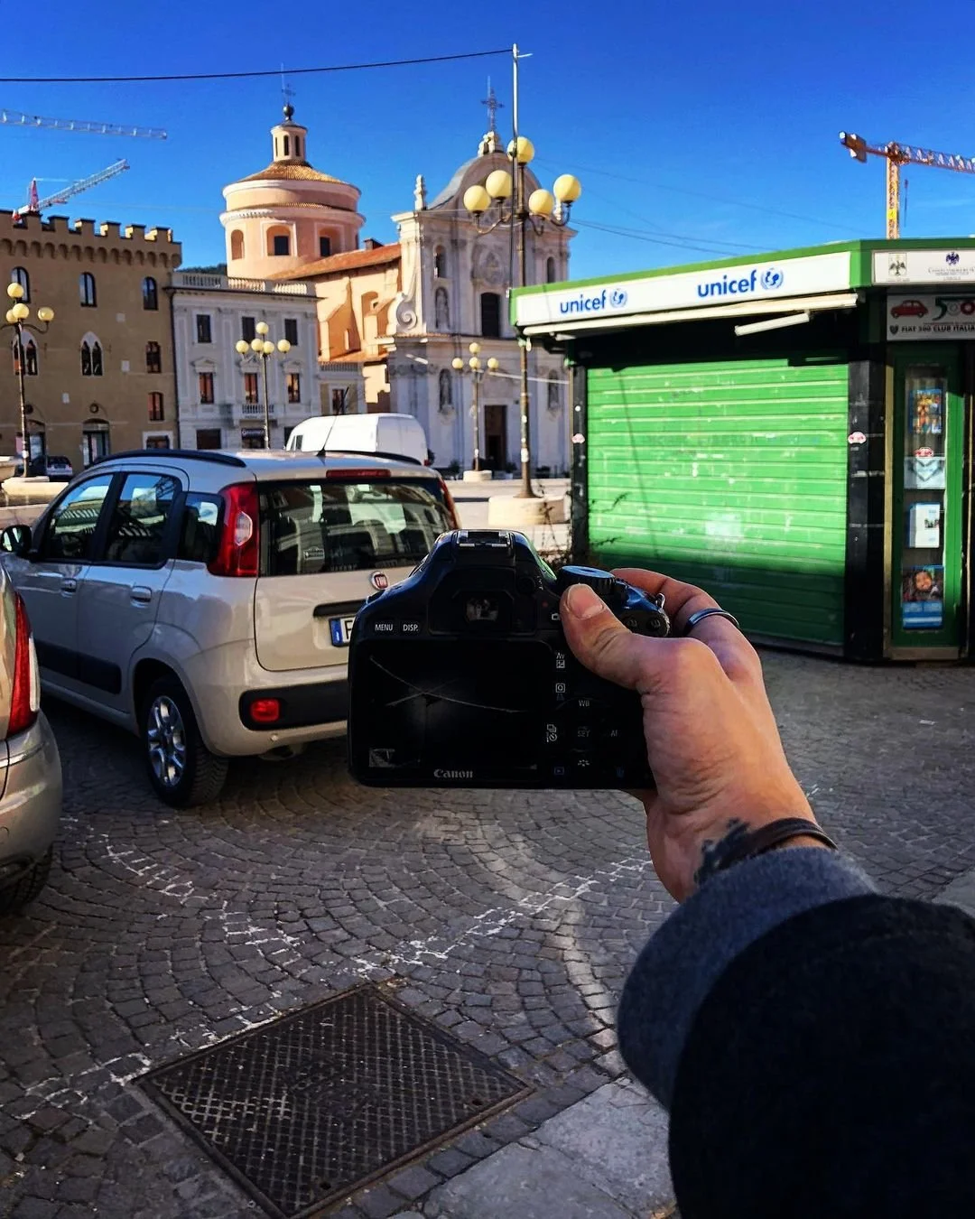 Person holding a Canon camera in a parking lot, capturing a cityscape with historical buildings, parked cars, and a green UNICEF kiosk against a clear blue sky.