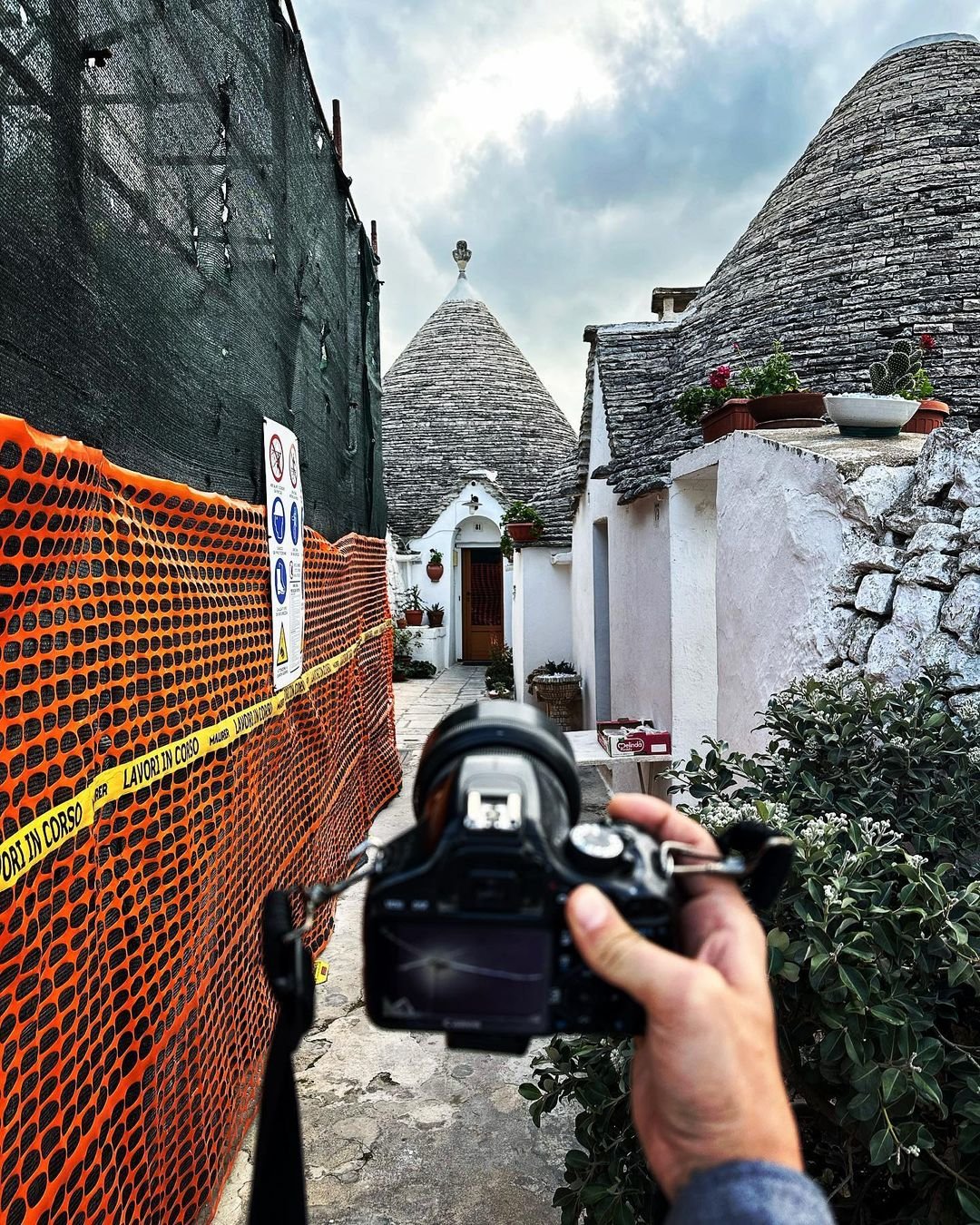 A person holding a camera in front of a pathway with construction fencing on the left and traditional white buildings with cone-shaped roofs on the right, under a cloudy sky.