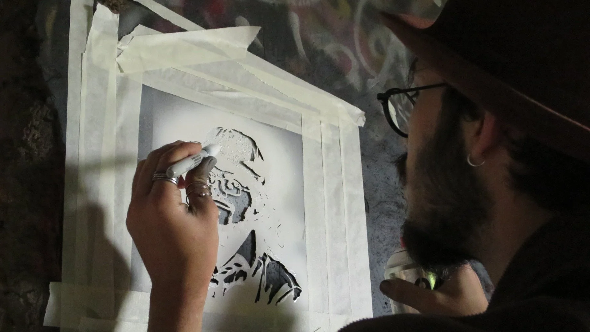 A person is creating a stencil art piece of a man's face on paper using a white marker. The person is wearing glasses and a hat, and the artwork is taped to a surface.