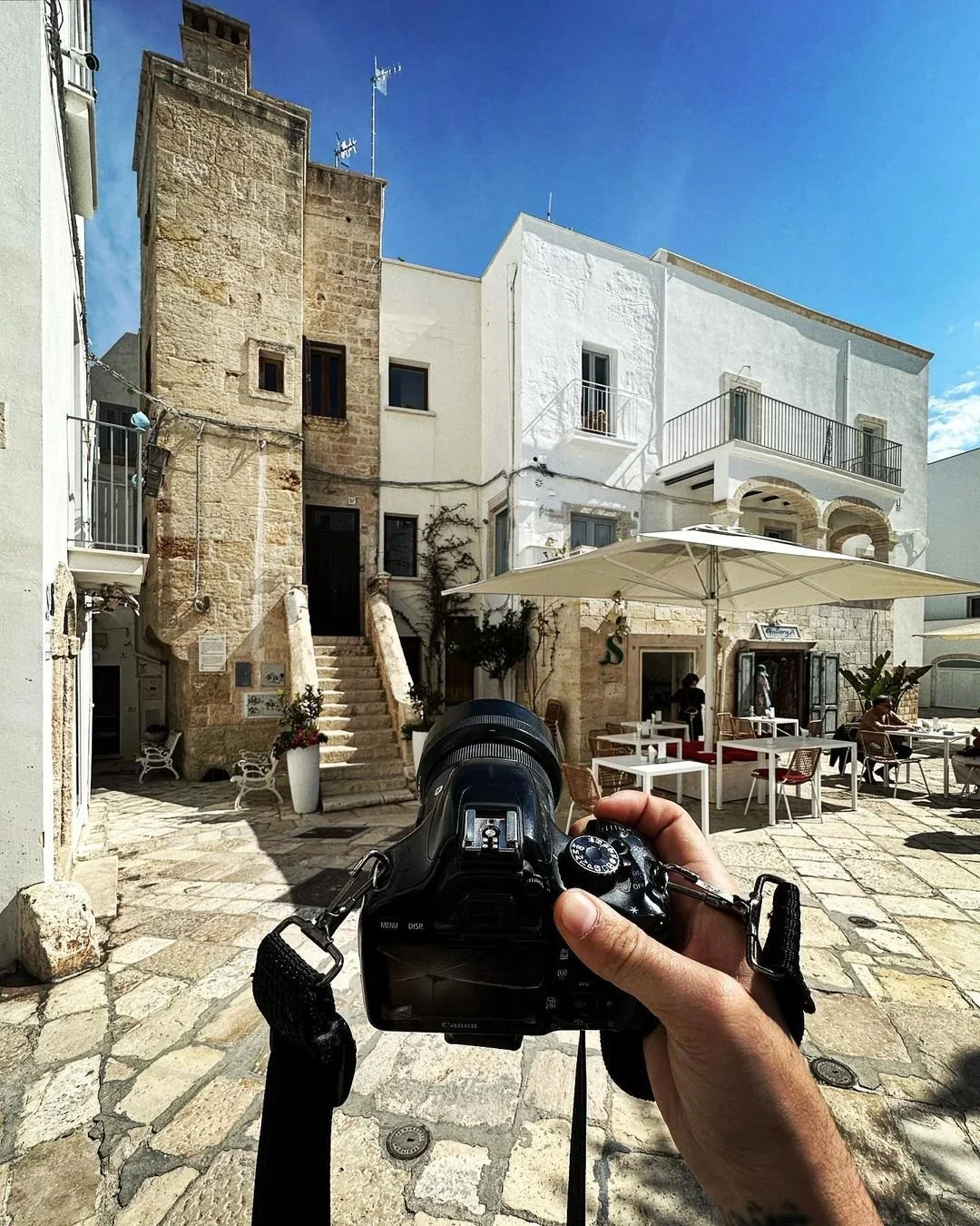 Person taking a photo of white and stone buildings with outdoor seating under a large umbrella, in a sunny courtyard with cobblestone pavement.