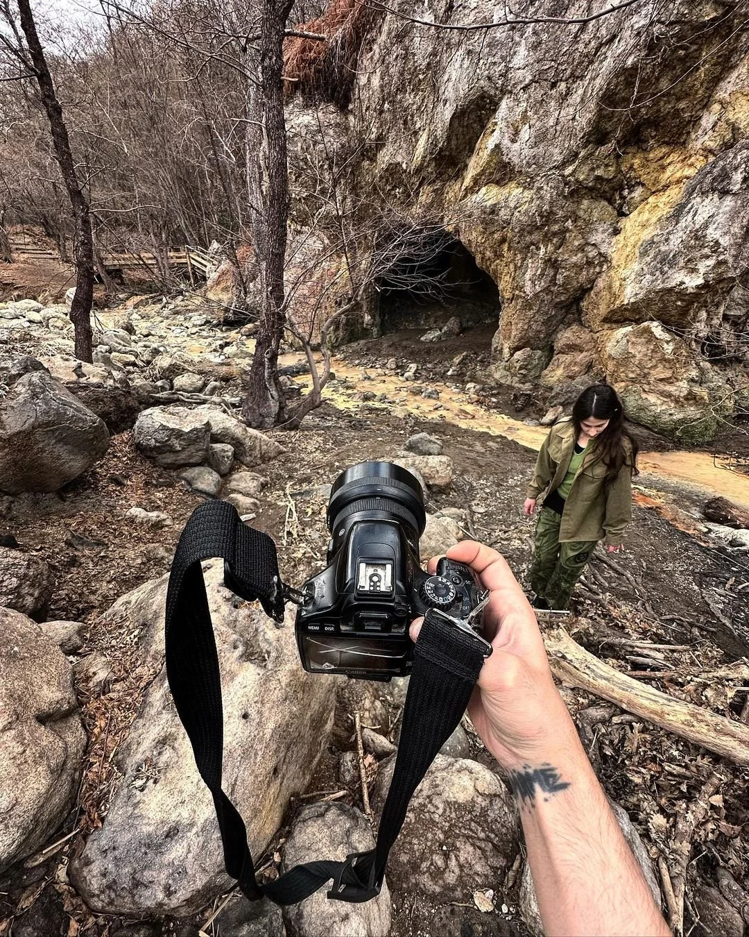 A person holding a camera takes a photo of a woman standing on a rocky, wooded trail near a cave entrance in a forest during late fall or early winter.