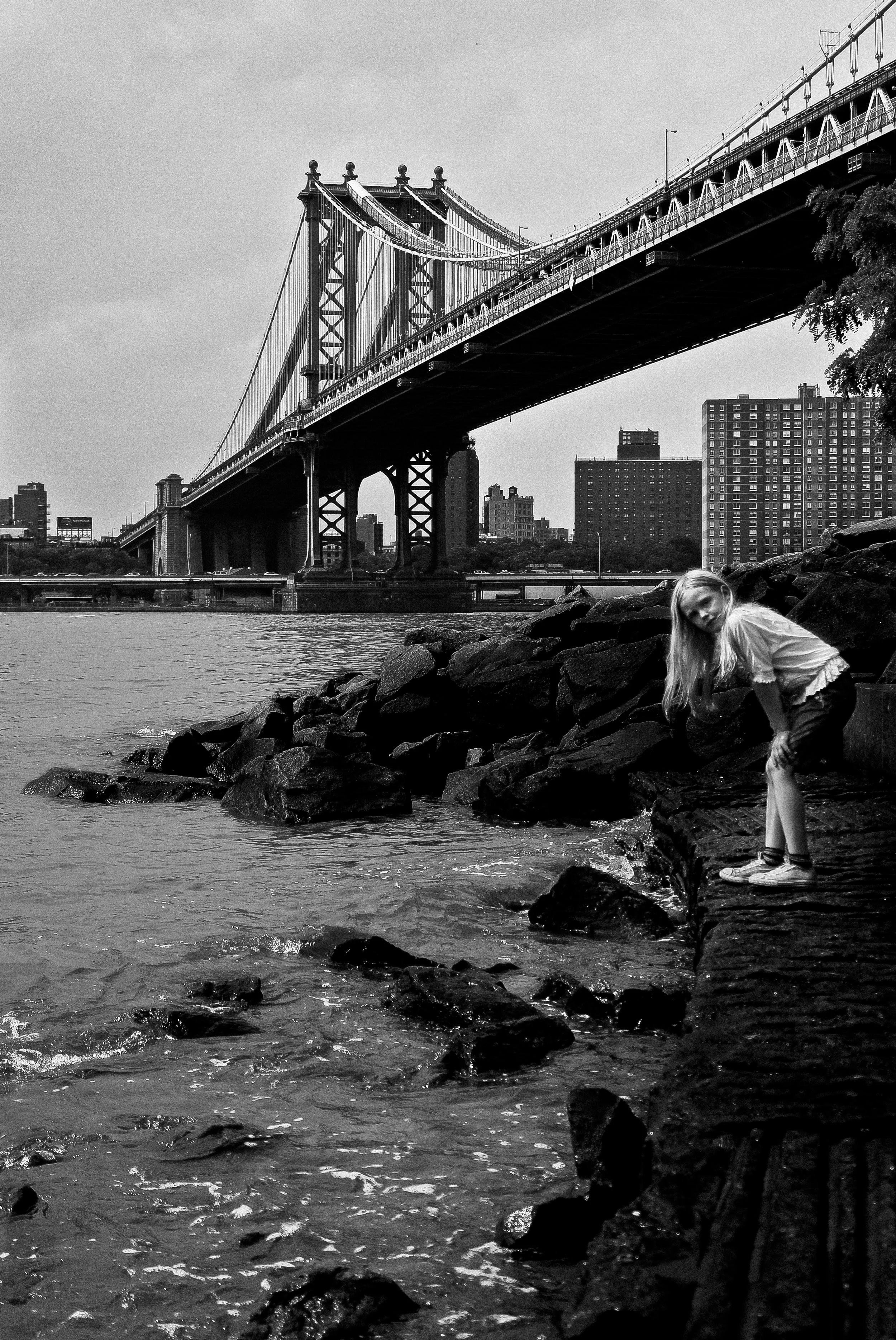 A black-and-white photo of a suspension bridge over a river, with a cityscape in the background. In the foreground, a young girl with long hair stands on rocks by the water, looking at the camera.