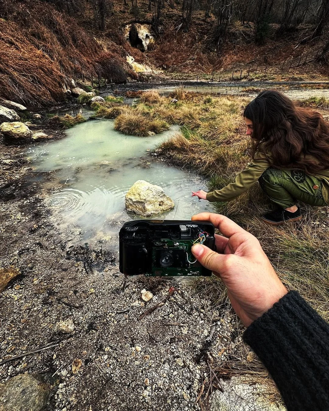 A person is crouching near a small pond in a natural outdoor setting, with their hand reaching toward the water, while another person is holding a camera pointed at the scene.