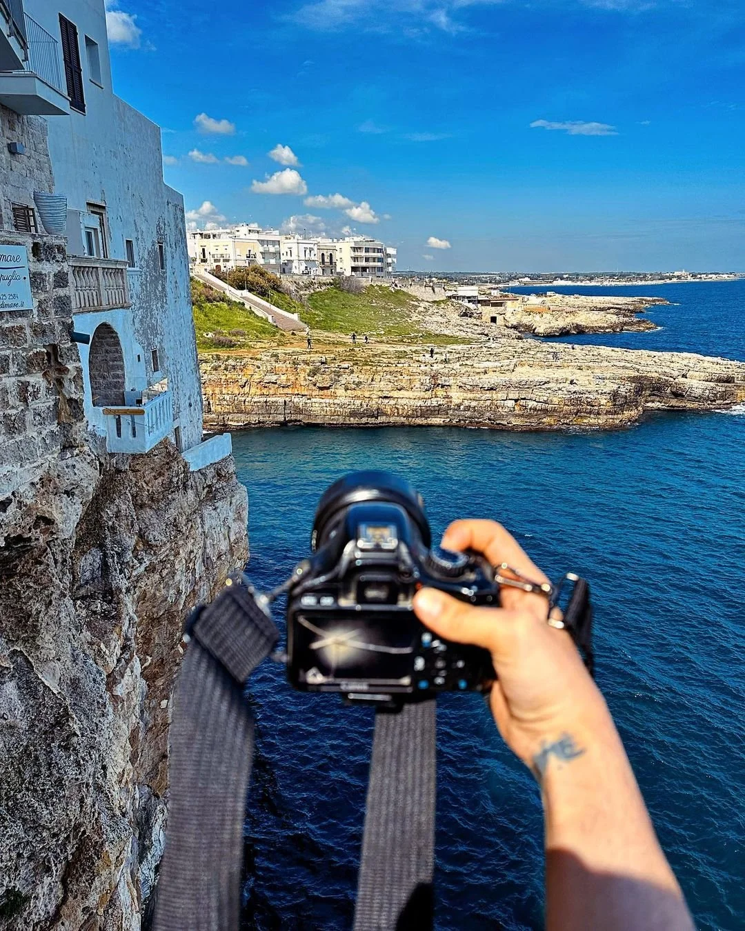 Person taking a photograph of a coastal scene with a DSLR camera, overlooking rocky cliffs, blue waters, and white buildings under a partly cloudy sky.