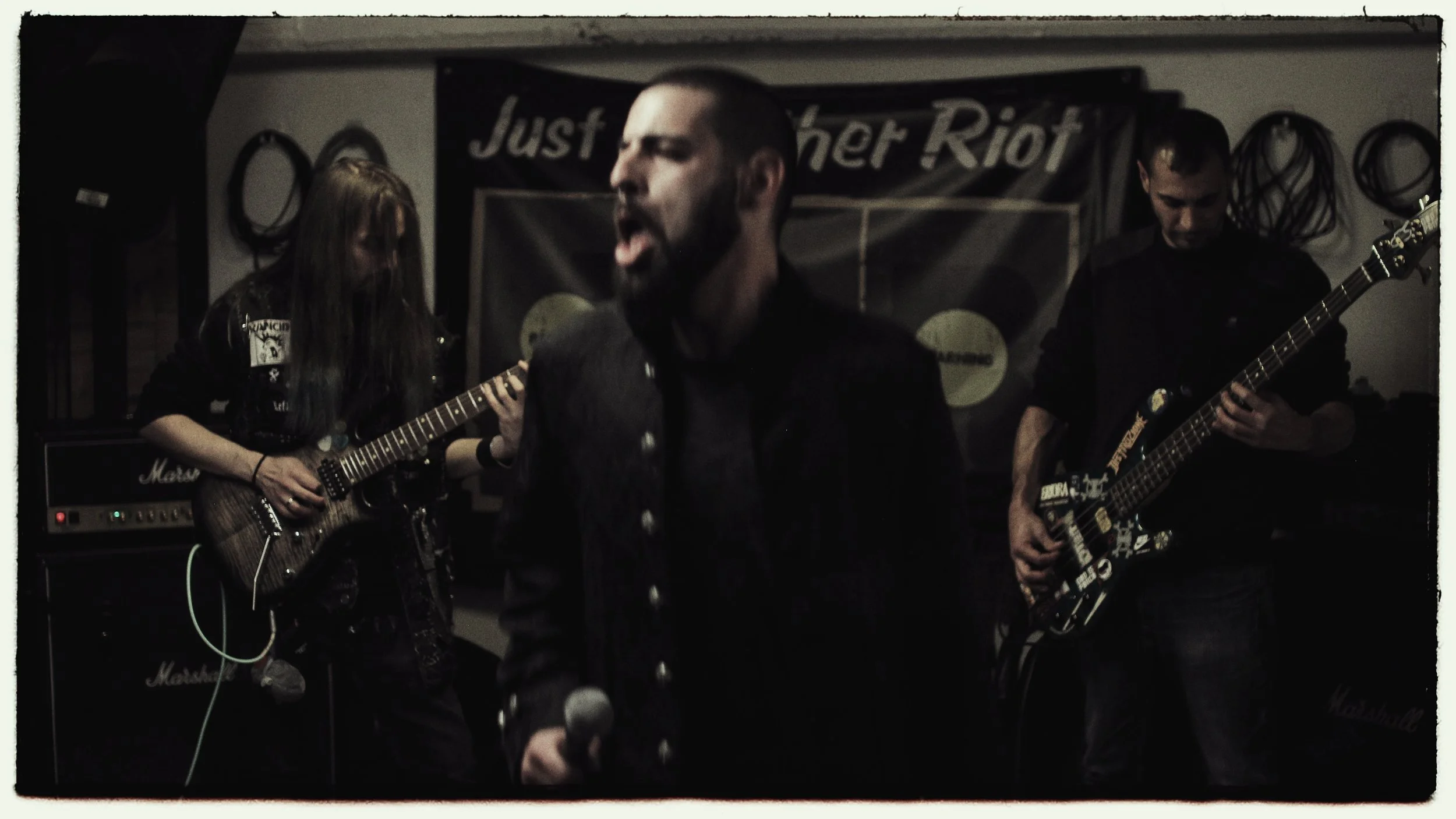 A band performing live with a vocalist singing into a microphone, a female guitarist on the left, and a male bassist on the right, in a dimly lit room with a banner reading 'Just Another Riot' in the background.