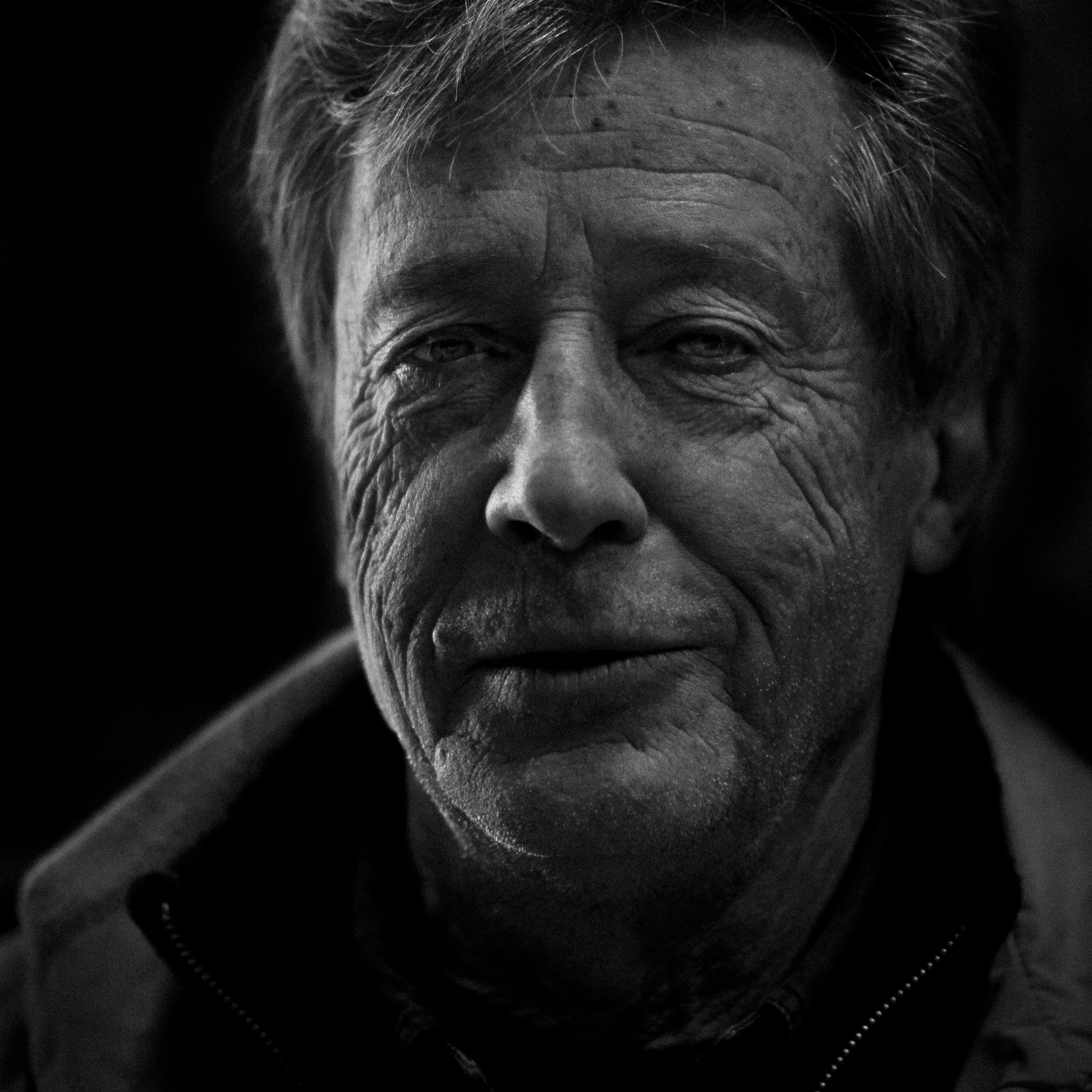 Close-up of an elderly person's face with deep wrinkles, gray hair, and a contemplative expression, captured in black and white.