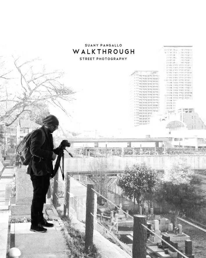 Black and white photo of a person standing on a sidewalk with a camera, looking at the view of city buildings and trees.