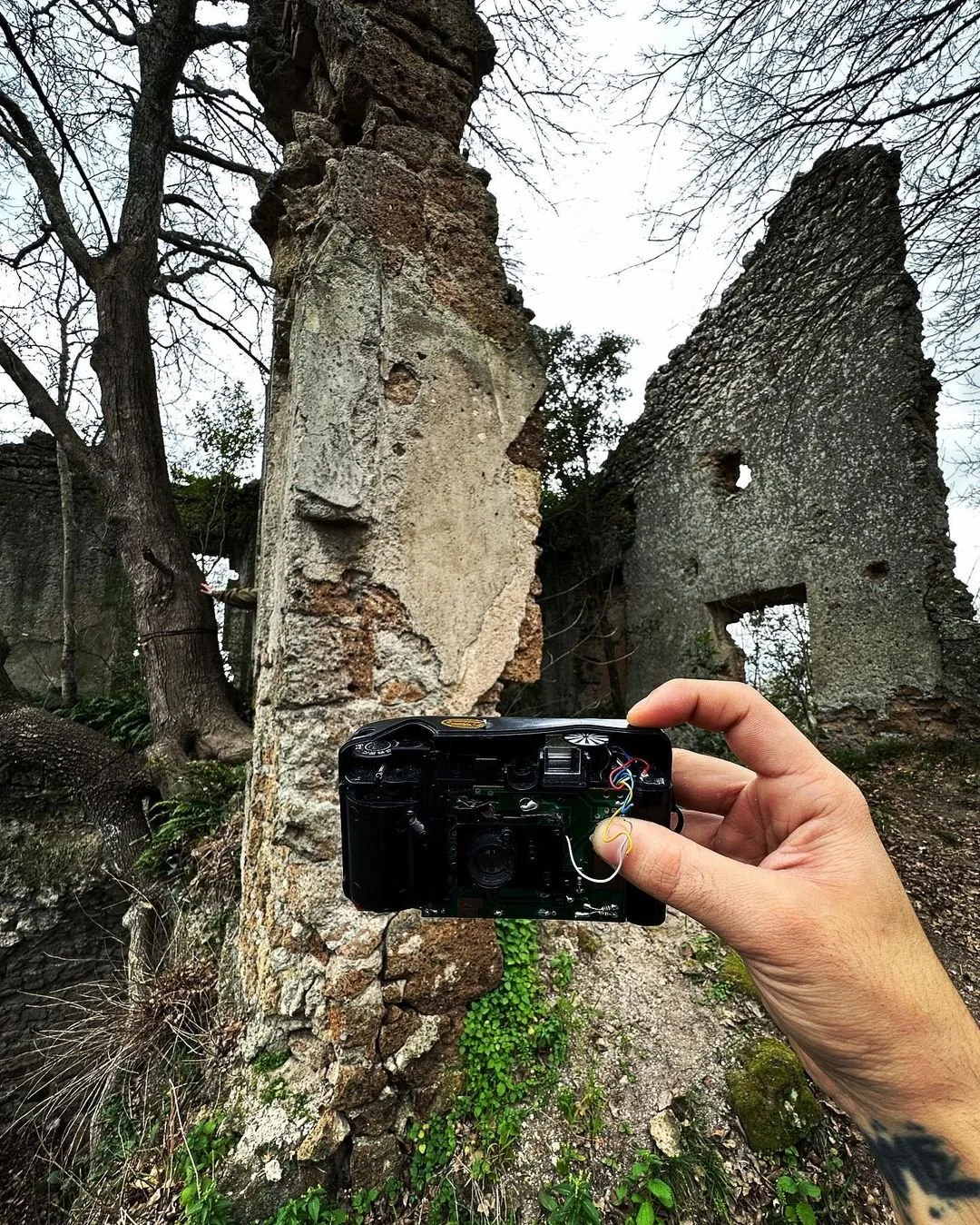 A person holds an old camera in front of the ruins of a stone building with broken walls and leafless trees.
