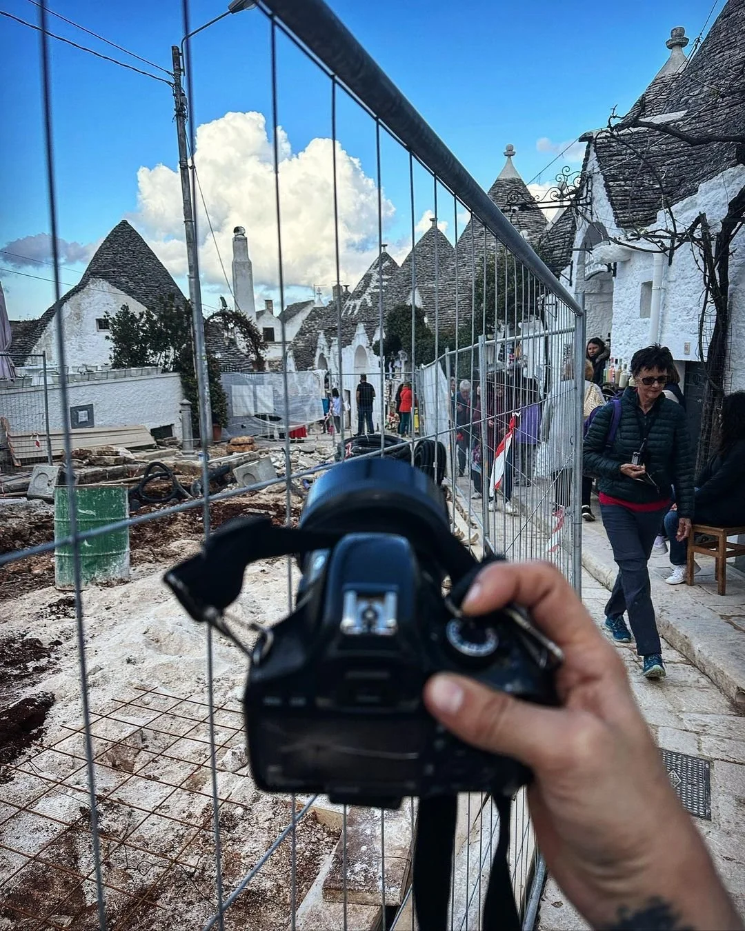 A person holding a camera takes a photo of tourists walking past a construction site with a wire fence. The background features white buildings with conical rooftops on a cloudy day.